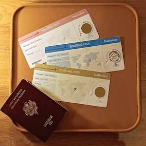 Puede incluir: Un conjunto de elementos de viaje sobre una bandeja de madera. Incluye tres tarjetas de embarque en rosa, azul y beige, y un pasaporte burdeos. Las tarjetas de embarque presentan un mapa del mundo, un avión y un diseño de globo.