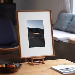 Puede incluir: Un marco de madera con un paspartú blanco que contiene una fotografía de paisaje. La foto muestra una oscura cordillera bajo un cielo azul claro con un lago naranja. El marco está sobre un caballete de madera.