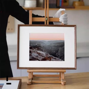Peut inclure: Une photographie encadrée d'un paysage montagneux avec un coucher de soleil rose et orange en arrière-plan. Le cadre est en bois foncé et est posé sur un chevalet en bois.