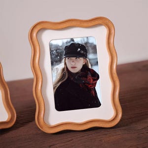 May include: A wooden picture frame with a wavy edge. The frame has a light brown finish and a white interior border. The frame holds a photograph of a person wearing a black hat and coat. The photo is set against a snowy background.