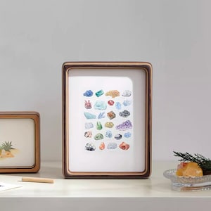 May include: A framed print featuring a watercolor illustration of various colorful gemstones. The frame is a warm brown color, and the print is displayed on a white background. The gemstones are in shades of blue, green, purple, and orange.