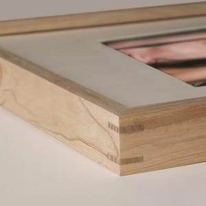 Puede incluir: Primer plano de un marco de fotos de madera con un acabado de madera clara. El marco tiene un diseño limpio y moderno con juntas de cola de milano visibles en las esquinas. El marco está hecho de madera en capas y tiene un passe-partout blanco.