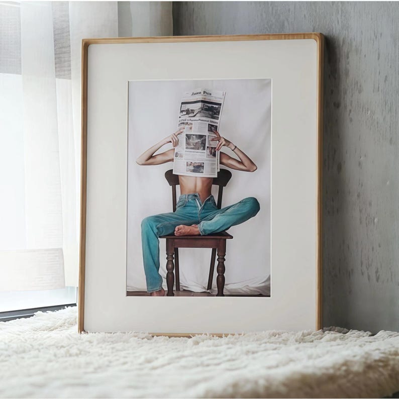 A large vertical white ash picture frame with a wide white mat, leaning beside a window and displaying artwork of a seated person reading a newspaper.