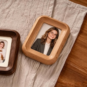 May include: Two wooden picture frames, one light-colored and one dark-colored, sit on a neutral-toned surface. The light-colored frame holds a photo of a person wearing glasses and a blazer. The dark-colored frame holds a photo of a person holding a pet.