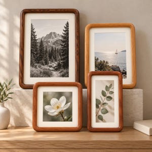 May include: Four wooden picture frames with rounded corners and white matting. The frames display a black and white mountain forest scene, a coastal sailboat view, a single white flower, and a green leafy branch on a light-colored surface.
