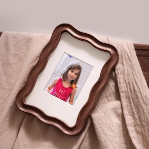 May include: A wooden picture frame with a wavy edge, holding a photograph of a young girl. The frame is a dark brown color, and the photo has a white mat. The girl is wearing a pink tank top with the words "DAY DREAMS".