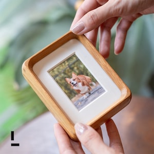May include: A wooden picture frame with a light-colored wood finish. The frame holds a photograph of a corgi dog running outdoors. The frame has a white mat and rounded corners.