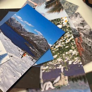 May include: A collection of printed photographs featuring various scenic landscapes. Images include a mountain range with snow, a blue lake, a field of daisies, and a palm tree. The photographs are arranged in a fan-like pattern.