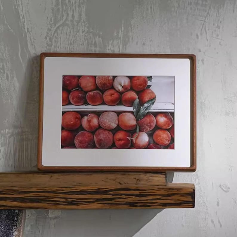 A horizontal teak picture frame with rounded corners and a white mat, displaying a red fruit still-life print while resting on a rustic wooden wall shelf.