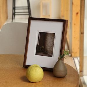 Può includere: Una cornice in legno marrone scuro con un passe-partout bianco, che mostra una fotografia di una stanza con una sedia. Un piccolo vaso di ceramica marrone con fiori bianchi e una pera gialla sono su un tavolo di legno.