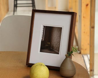 Könnte beinhalten: Ein dunkelbrauner Holzbilderrahmen mit weißem Passepartout, der ein Foto eines Raumes mit einem Stuhl zeigt. Eine kleine, braune Keramikvase mit weißen Blumen und eine gelbe Birne stehen auf einem Holztisch.