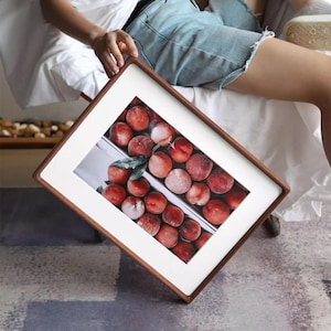 A seated person holds a large white ash picture frame on their lap, displaying a red fruit still-life print with a wide white mat.