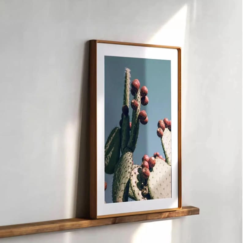 A tall vertical solid wood picture frame with softly rounded corners, displaying a cactus print with red fruit and leaning on a narrow wooden ledge against a white wall.