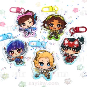 May include: Five acrylic keychains featuring cartoon characters with various hair colors and outfits. Each keychain has a different character design and a colored clasp. The keychains are on a white surface with star-shaped confetti.