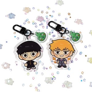 Könnte beinhalten: Zwei Schlüsselanhänger aus Acryl mit Anime-Charakteren. Ein Schlüsselanhänger zeigt einen Charakter mit schwarzen Haaren und Schuluniform, der andere mit orangefarbenen Haaren und Anzug. Jeder Schlüsselanhänger hat einen schwarzen Verschluss und einen grünen geisterhaften Anhänger.