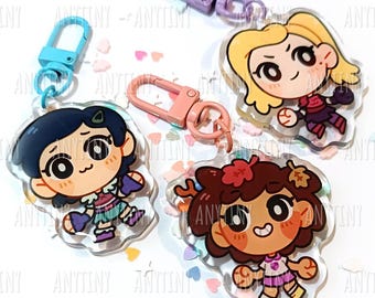 May include: Four cartoon character keychains with colorful clasps. The keychains feature three different characters with large eyes and smiling faces. The characters are wearing different outfits. The keychains are on a white surface with heart-shaped confetti.