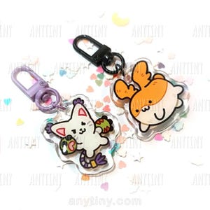 May include: Two clear acrylic keychains with cartoon animal designs. One features a white cat with red and green accents, the other an orange and white creature. Both have black metal clasps and are surrounded by colorful confetti.