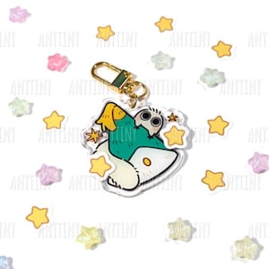 Può includere: Un portachiavi in acrilico trasparente con un design a gufo dei cartoni animati. Il gufo ha un corpo verde, ali gialle e pancia bianca. Ha un fermaglio color oro ed è circondato da decorazioni a forma di stella gialle e pastello.