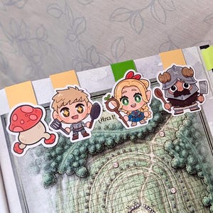 Puede incluir: Una colección de marcadores con personajes coloridos. Los marcadores presentan ilustraciones de un hongo, un caballero con espátula y cuchara, una mujer con un bastón y un guerrero con una cuchara. Los marcadores están colocados en un libro con un mapa.