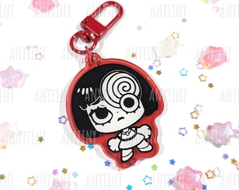 May include: A red keychain featuring a cartoon character with black hair and a spiral design on the head. The character is outlined in white and has a sad expression. The keychain has a red clasp and the website address anytiny.com is visible.