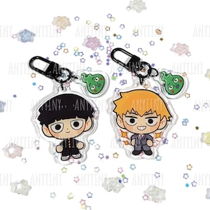 Puede incluir: Dos llaveros de personajes de estilo anime. Un llavero presenta un personaje con cabello negro y uniforme escolar, el otro con cabello naranja y un traje. Cada llavero tiene un amuleto de espíritu verde y un cierre negro.