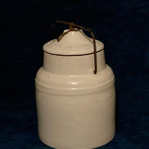 May include: A white ceramic jar with a lid and a wire handle. The jar has a cylindrical body and a slightly tapered top. The lid has a raised center and a decorative rim. The wire handle is attached to the lid and the jar.