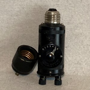 May include: A black vintage light bulb socket with a numbered dial and a detached base. The socket has a silver-coloured screw-in base and two small black knobs at the bottom. The dial is marked from 0 to 6.