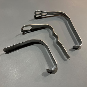 May include: Three stainless steel medical instruments are arranged on a gray surface. The instruments have curved, elongated shapes with various end designs, including a slotted tip and a ridged edge. The tools are likely used in medical procedures.
