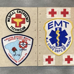 Könnte beinhalten: Eine Sammlung von gestickten Aufnähern und Emblemen im Zusammenhang mit dem Rettungsdienst. Enthält Aufnäher mit Texten wie "Medical Emergency Technician", "EMT" und "Lancaster Emergency Medical Services Association". Die Aufnäher zeigen rote Kreuze und medizinische Symbole.