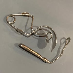 May include: A silver metal dental retainer with a curved wire design and a gold-colored section. The retainer is designed to fit in the mouth and is likely used to maintain the position of teeth after orthodontic treatment.