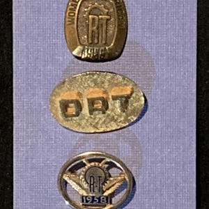 May include: Three vintage metal pins on a lavender background. The top pin is oval with the text "WOMEN'S AMERICAN RT 1949". The middle pin is oval with "DAT" on it. The bottom pin is round with "RT 1958" and wings.