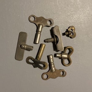 May include: A collection of six antique clock winding keys. The keys are made of metal and have different shapes and sizes. Some of the keys have a decorative design.