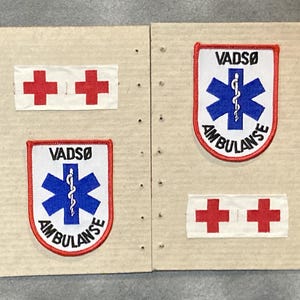 May include: Two sets of medical patches and two rectangular patches with red crosses. The patches feature the text "VADSO AMBULANSE" and a blue star of life symbol. The patches are on a cardboard background.
