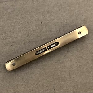 Puede incluir: Un nivel de latón rectangular con dos pequeños viales de burbujas. El nivel tiene un acabado pulido y dos pequeños agujeros en cada extremo. Está diseñado para medir superficies horizontales y verticales.