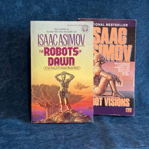 May include: Two science fiction paperback books by Isaac Asimov. The book on the left is "The Robots of Dawn" and the book on the right is "Robot Visions".
