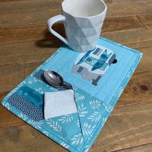 May include: A blue and white fabric coaster with a camper design and a white mug on top. The coaster has a pocket for a spoon and a napkin.