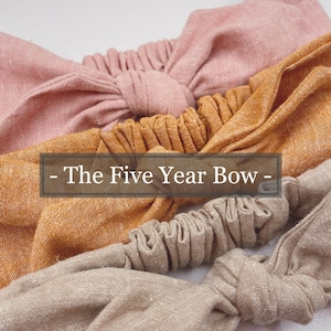 May include: Three fabric hair scrunchies in different colors: pink, mustard yellow, and beige. The scrunchies are tied together in a bow and the text "The Five Year Bow" is printed on a black banner.
