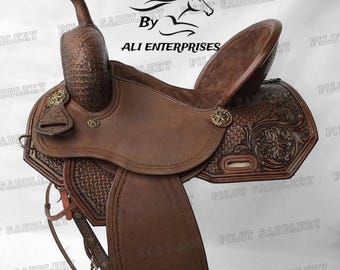 May include: A brown leather saddle with intricate tooling and a tooled floral design. The saddle is made by Pilot Saddlery, Ali Enterprises.