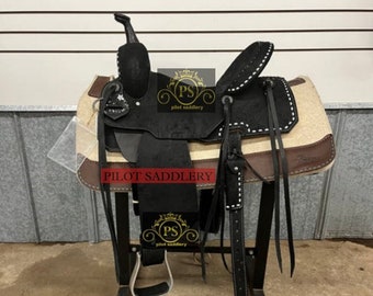 May include: A black leather saddle with a brown leather seat and white fleece padding. The saddle is on a black metal stand. The saddle has a silver concho and a black leather stirrup. The saddle is labeled "Pilot Saddlery".