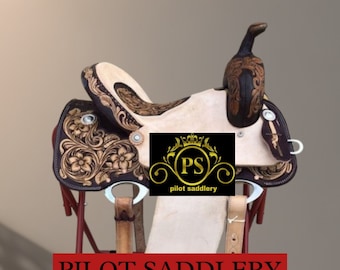 May include: A brown leather saddle with intricate floral tooling. The saddle is on a red metal stand and includes a stirrup and a leather cinch. The saddle is labeled "Pilot Saddlery".
