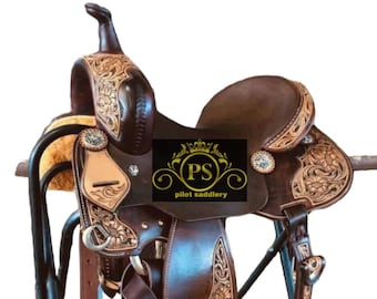 May include: A brown leather western saddle with floral tooling and decorative silver accents. The saddle is displayed on a black metal stand. The text "PS pilot saddlery" is visible on the saddle.