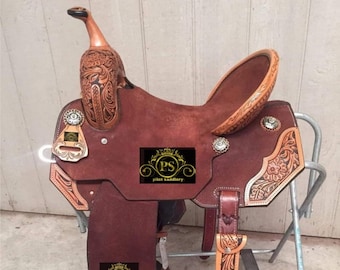 May include: A brown leather saddle with intricate tooling and floral designs. The saddle features a  suede seat, silver conchos, and a  'Pilot Saddlery' logo.