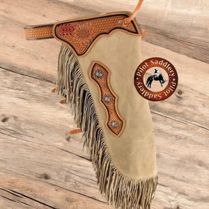 Può includere: Chaps in pelle scamosciata beige con lavorazione decorativa e frange. Gli chaps presentano un motivo a girasole e borchie decorative in metallo. Il logo Pilot Saddlery è visibile. Progettato per essere indossato attorno alle gambe e alla vita.