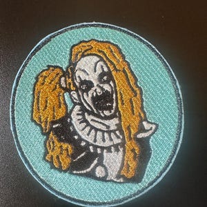 Può includere: Un cerotto rotondo ricamato di colore blu con un contorno bianco. Il cerotto presenta un clown spaventoso con capelli lunghi e arancioni e un colletto bianco. Il clown sorride con un ampio sorriso pieno di denti.