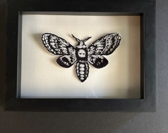 May include: A black and white embroidered moth with a skull-like pattern on its wings is framed in a black frame.
