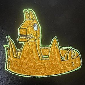 May include: A brown embroidered patch depicting a cartoon llama wearing a crown. The llama is sitting in a boat with a green outline.