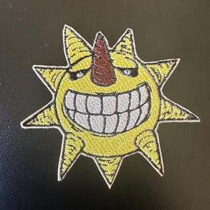 May include: A yellow embroidered patch of a cartoon sun with a large toothy grin and a brown nose. The sun has a spiky outline.