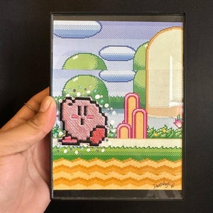 May include: A framed pixel art piece featuring the character Kirby in a colorful landscape. The artwork includes green hills, blue skies with clouds, and pink and yellow elements. The artist's signature is visible in the bottom right corner.