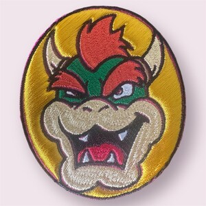 Peut inclure: Patch brodé représentant un gros plan du visage de Bowser. Bowser est un personnage fictif de la série de jeux vidéo Super Mario. Il a une carapace verte et hérissée, des cheveux roux et une expression menaçante. Le patch est circulaire et a un fond jaune.