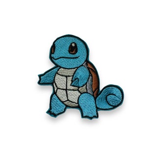 Puede incluir: Un parche bordado con una tortuga de dibujos animados con caparazón. La tortuga es azul y marrón y camina con la cabeza girada hacia un lado.